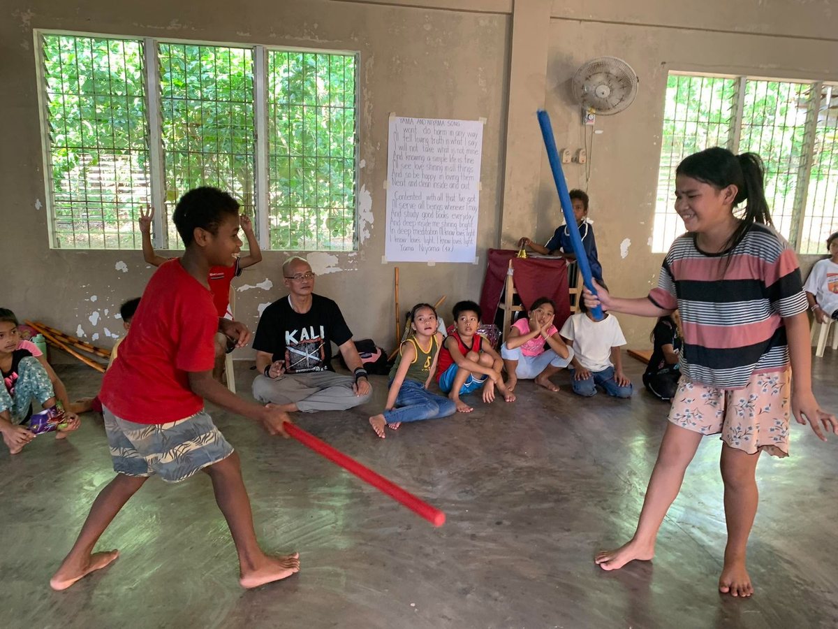 Two children spar with padded sticks in an Arnis class at AMSAI School while classmates and their instructor watch