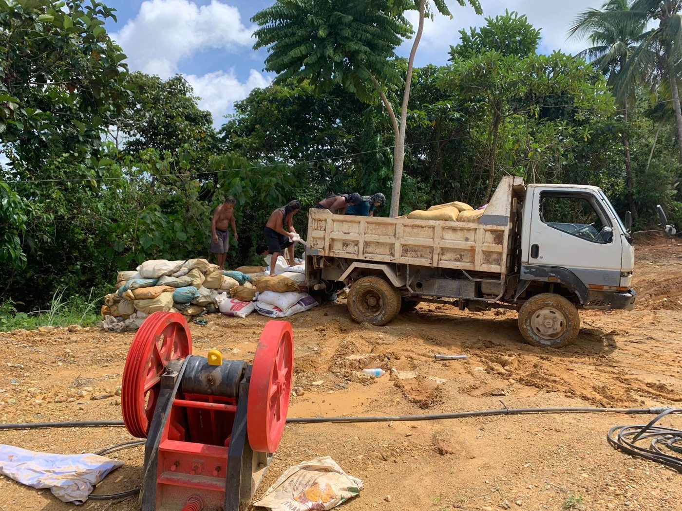 Workers load bags of ore onto a dump truck, with a red jaw crusher in the foreground